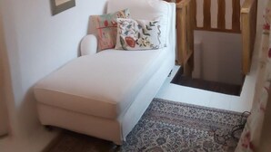 Une petite pièce accueillante, avec un canapé blanc, un banc en bois et un tapis à motifs. Plusieurs tableaux encadrés ornent le mur, et une fenêtre offre une vue sur la verdure.