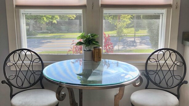 Spiseområde med et rundt glasbord og to stole, en potteplante og udsigt til en gade gennem et vindue med persienner.