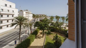 Une vue sur la côte, depuis un balcon donnant sur une plage de sable bordée de palmiers, avec une route et des bâtiments modernes en arrière-plan.