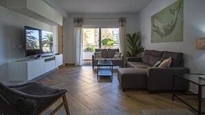 Un salon moderne doté d’un téléviseur à écran plat, d’un canapé, d’une table basse et d’une grande fenêtre offrant une vue sur les immeubles et les palmiers.
