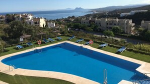 Una piscina con lettini, immersa nel verde e con edifici circostanti, con vista sul mare e montagne lontane.