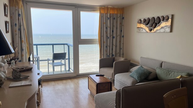 A living room with a sofa, a chair, a table, and a view of the sea through large windows. - Apartment with sea view (Bray-Dunes)