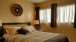 A neatly made bed with a plaid bedspread, a round wall clock, and a window with curtains. - Private Cozy Studio walking distance to beaches, shops, restaurants and bars (Kihei)