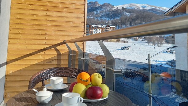 A balcony with a table set for two, featuring a fruit bowl and cups. A mountain landscape is visible in the background. - Bakuriani Crystal Loft 406 (Borjomi)