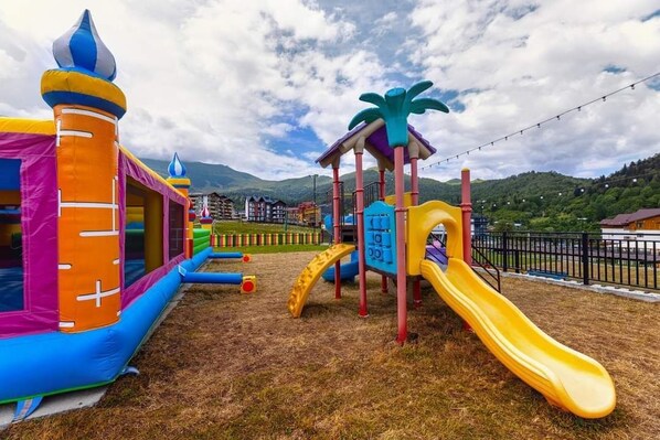 A playground with a slide and climbing structures, surrounded by a fence and buildings in the background. - Bakuriani Crystal Loft 406 (Borjomi)