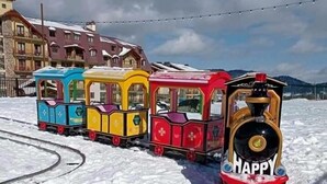 A snow-covered train track with colorful miniature trains, a 'HAPPY' sign, and buildings in the background. - Bakuriani Crystal Loft 406 (Borjomi)