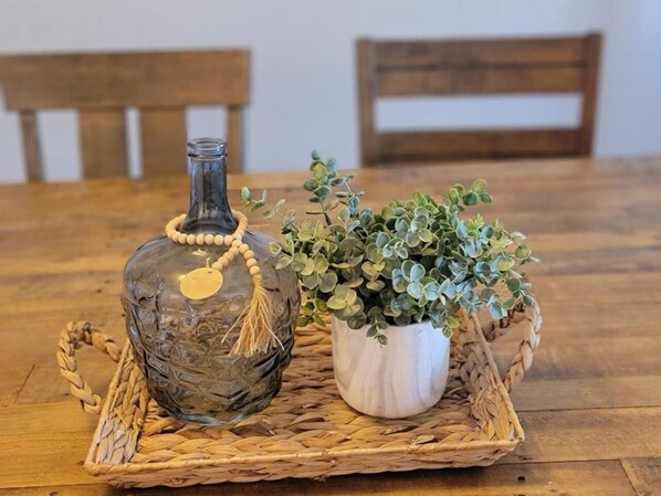 Une table en bois sur laquelle repose un plateau tressé. Sur ce plateau, on trouve une bouteille en verre avec une étiquette et une petite plante dans un pot blanc.
