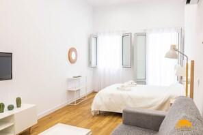 A modern hotel room with a bed, a grey sofa, a TV, and a small shelf with decorative items.
