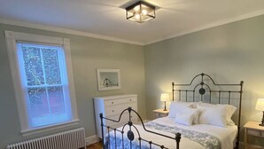 A bedroom with a metal bed frame, a bedside table with a lamp, a radiator, a window with blinds, and a patterned rug. - Saugerties Village Space with Great Backyard (Saugerties)