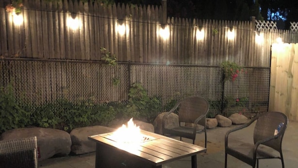 An outdoor patio area with a metal table and chairs, a fire feature, and a wooden fence with lights. - Saugerties Village Space with Great Backyard (Saugerties)
