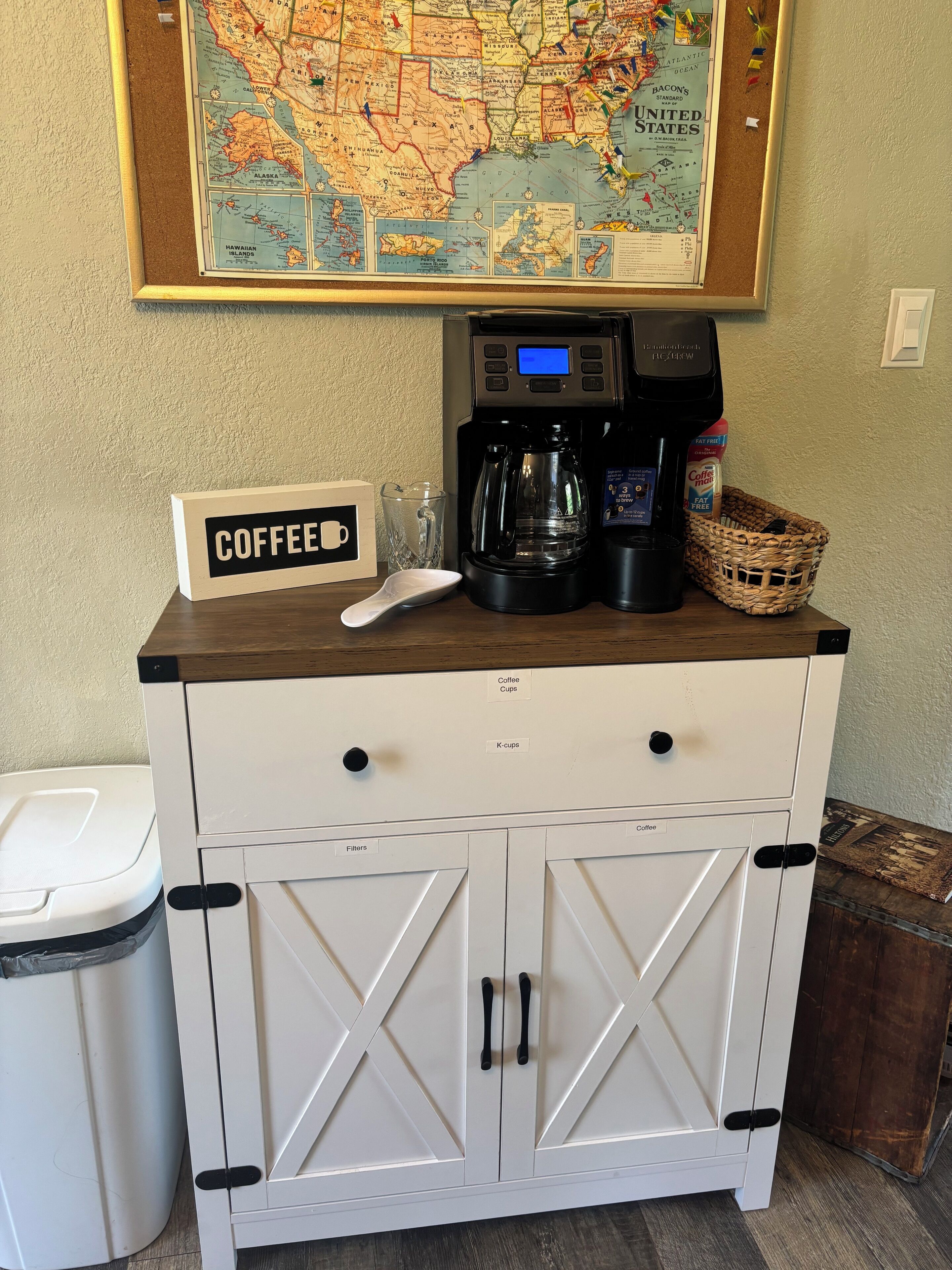 A coffee station with a sign, a coffee maker, and a basket of coffee pods.