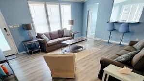 Un salón amplio con un sofá de cuero marrón, un otoman beige y una mesa de centro blanca. Hay dos taburetes azules y una lámpara sobre una mesita auxiliar.