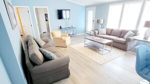 Una sala de estar moderna con un sofá gris, un sillón azul y una mesa de centro. Hay un televisor montado en la pared y un ventilador de techo.