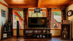 Una sala de estar acogedora con techo de madera, un televisor de pantalla plana grande, una estantería y una ventana con cortinas.