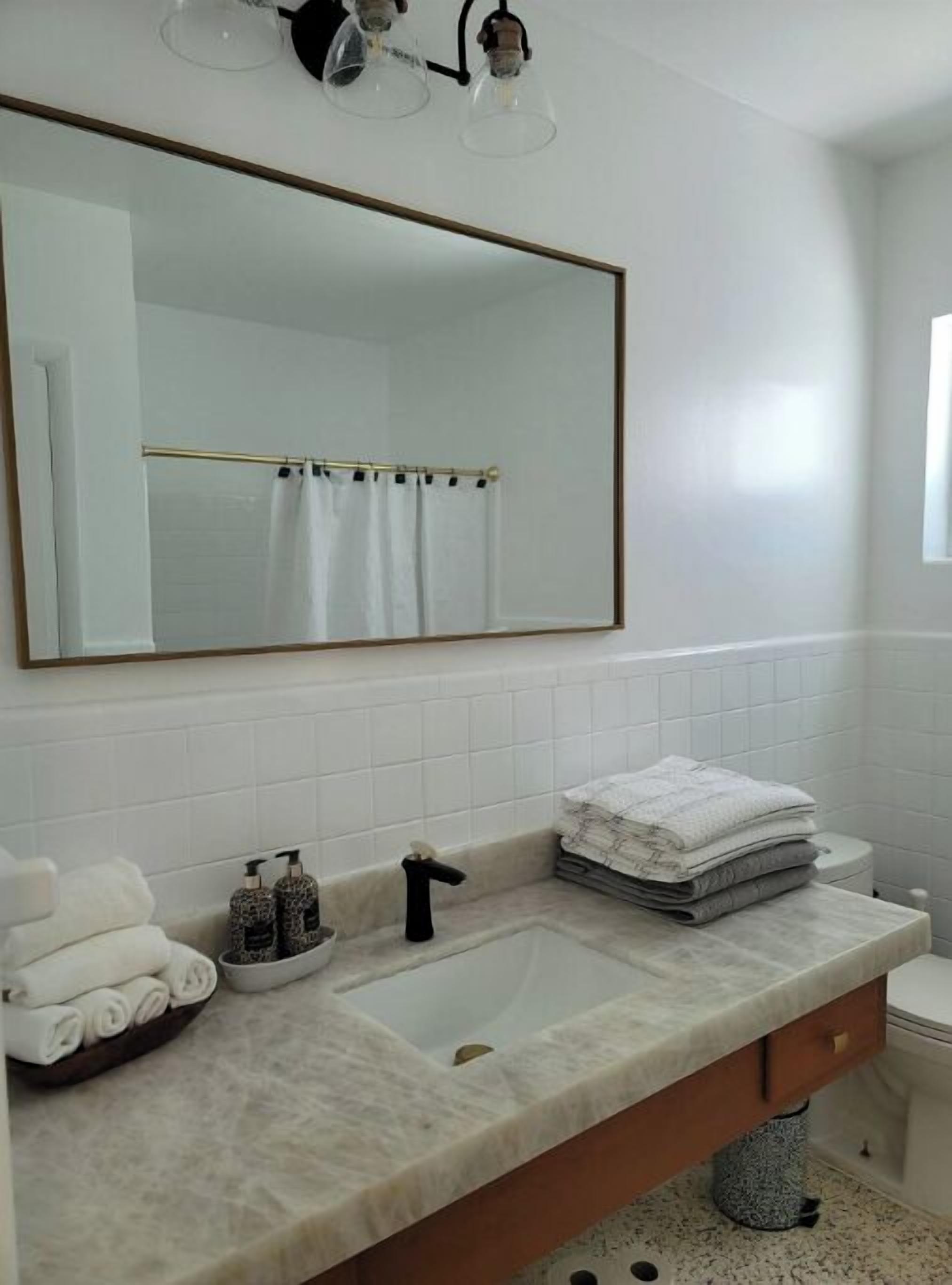 A bathroom with a large mirror, white tiles, a marble countertop, a sink, a toilet, and a towel rack with towels.