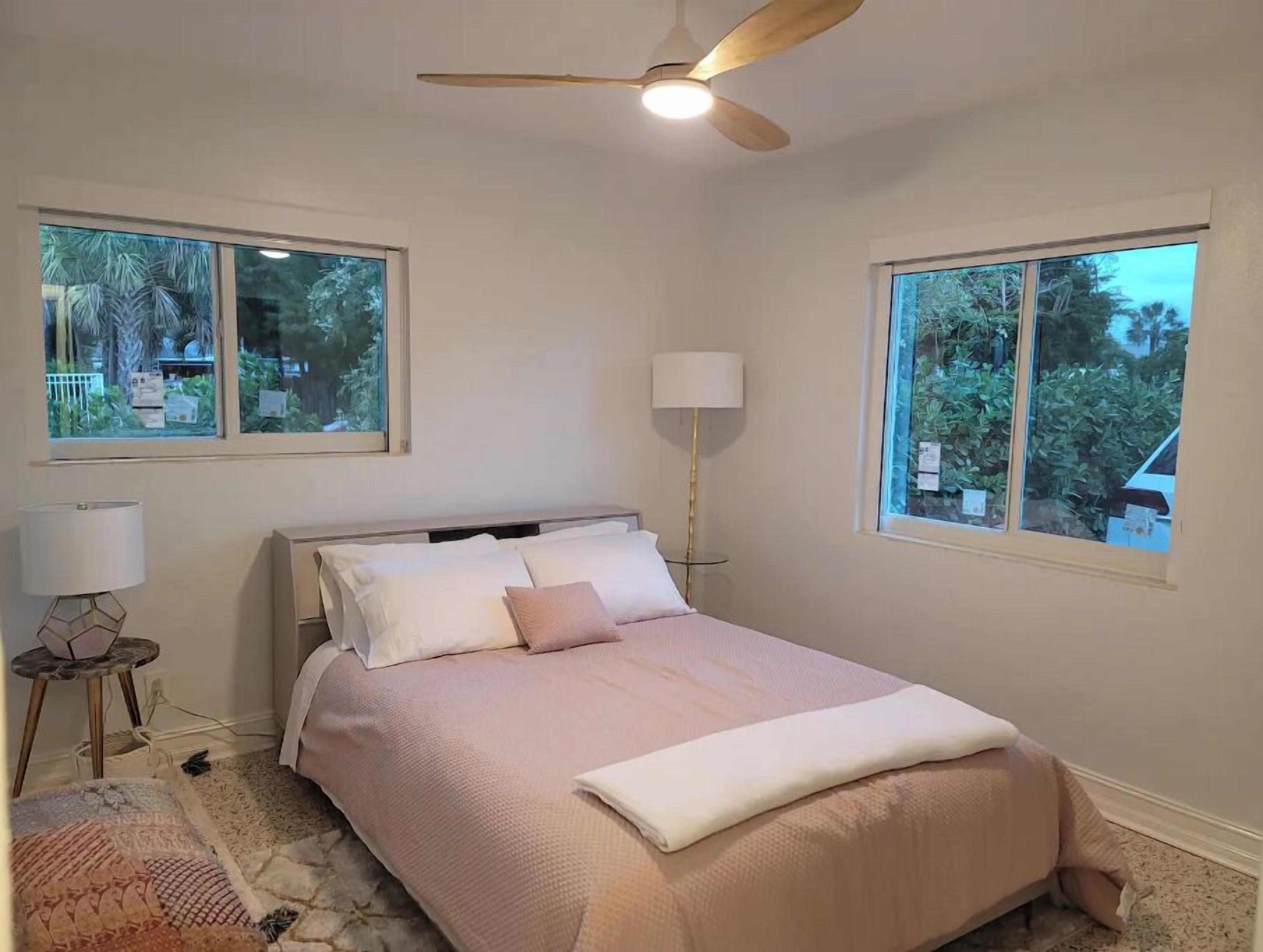 Une chambre à coucher avec un lit, deux fenêtres, un ventilateur de plafond et une lampe.