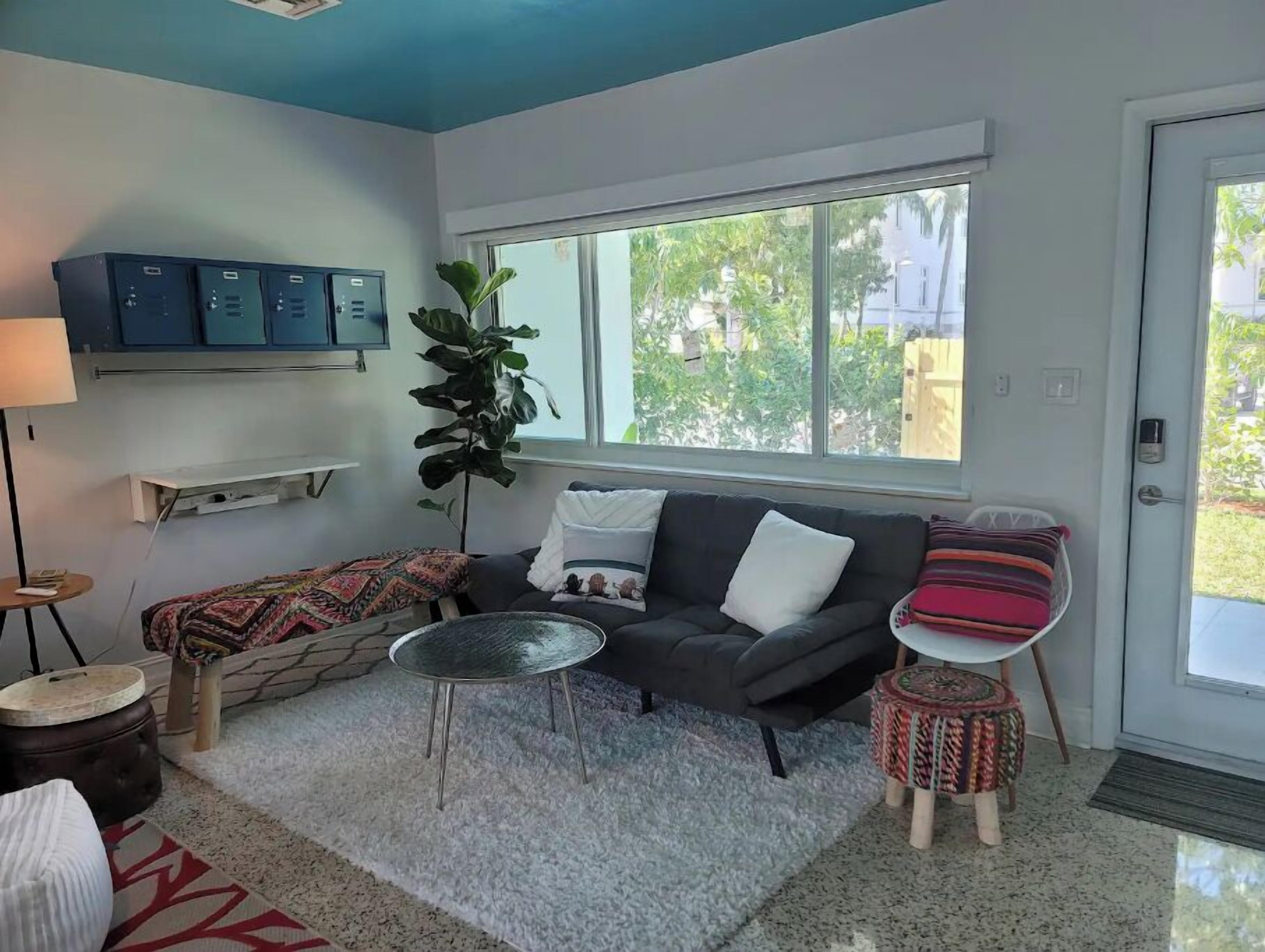 Un salon avec un canapé gris, une table basse ronde, un tapis à motifs, une plante en pot et une fenêtre donnant sur un paysage verdoyant.