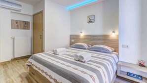 Habitación de hotel con cama, cabecero de madera, aire acondicionado de pared, radiador, puerta y mesita de noche con lámpara.