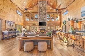 A spacious, high-ceilinged log cabin with a stone fireplace, a large dining table, bar stools, and a cozy seating area. - Sleeps 17 | 2 King Suites | Heated Pool & Hot Tub (Broken Bow)