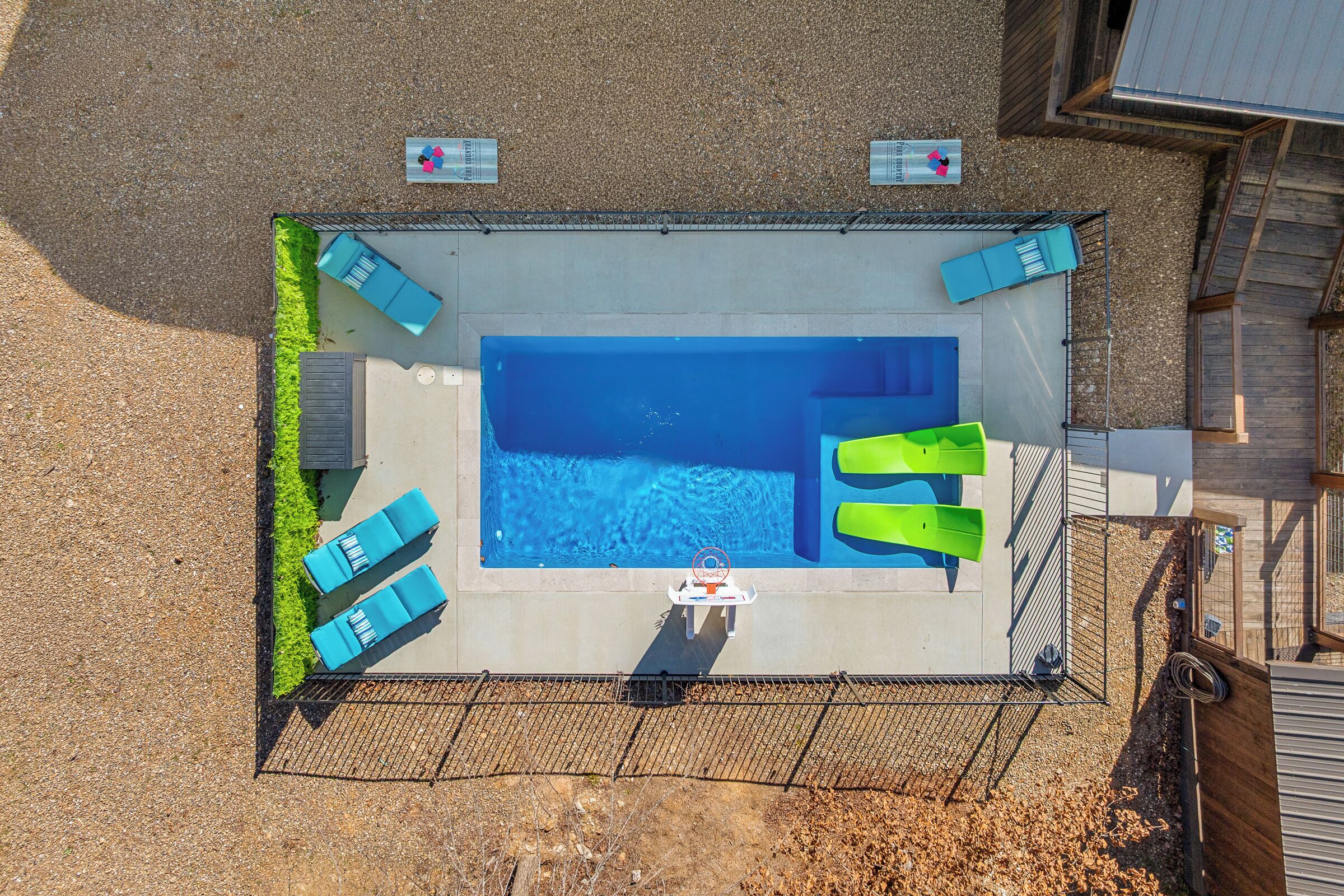 Eine Vogelperspektive auf einen rechteckigen Swimmingpool mit Sprungbrett, umgeben von einem Zaun und ausgestattet mit Liegestühlen.