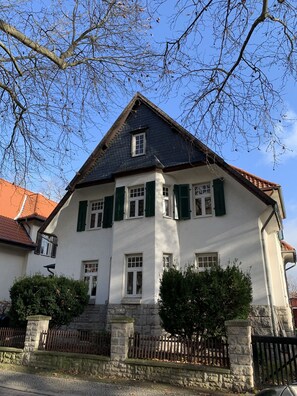 Une maison à deux étages, dotée d’un toit à forte pente, de murs extérieurs blancs et de volets verts.