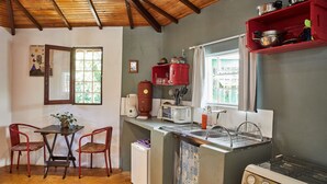 A compact kitchen with a wooden ceiling, a small dining table with chairs, a refrigerator, a stove, and a window with a view of greenery. - Chale Ipe Jatoba (Alto Paraíso de Goiás)