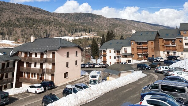 A parking lot with several cars and a snow-covered ground. There are buildings with multiple floors and balconies.