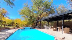 Piscina rectangular con un área de patio techada, rodeada de árboles y mobiliario de exterior.