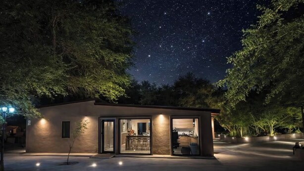 Un edificio moderno de un piso con entrada de vidrio, rodeado de árboles, bajo un cielo nocturno estrellado.