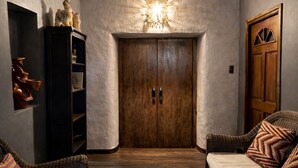 Una habitación con techo de madera, una puerta central, dos sofás de mimbre, un aparador con esculturas, y una lámpara fijada a la pared.