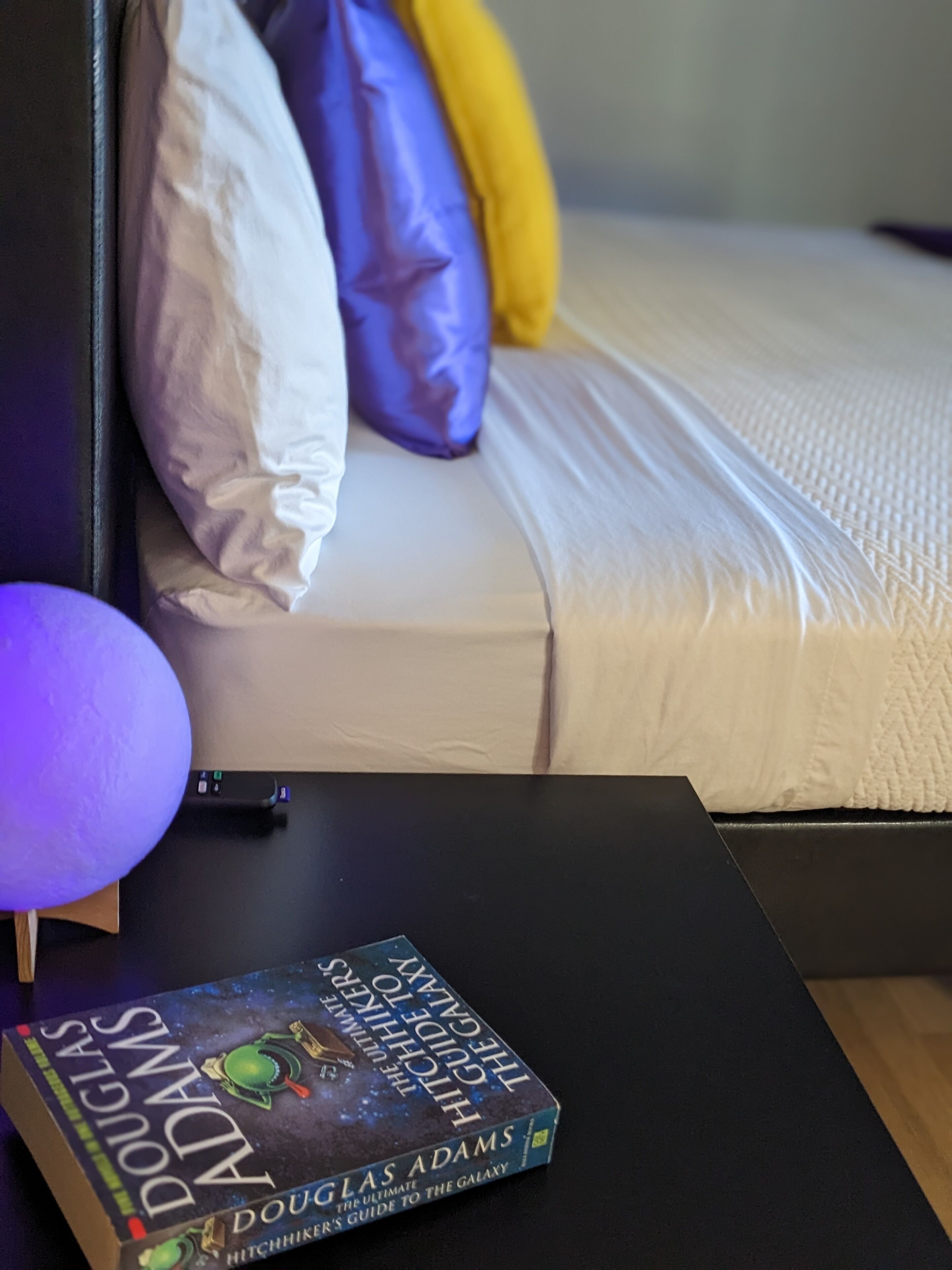 A neatly made bed with pillows, a bedside table with a book and a lamp, and a wall-mounted charging station.