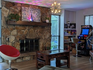 Una chimenea de piedra con un cuadro de un pavo real, una silla roja, una mesa de madera y un juego de arcade.
