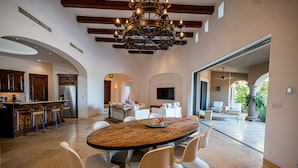 A spacious dining area with a wooden table and white chairs, a kitchen with dark wooden cabinets, and a large TV in the background. - Breathtaking Views, Private Pool/Spa adjacent to One&Only Palmilla!! (San José del Cabo)