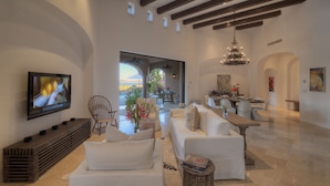 A spacious living area with a large flat-screen TV, white sofas, a wooden bench, and a dining table with chairs. - Breathtaking Views, Private Pool/Spa adjacent to One&Only Palmilla!! (San José del Cabo)