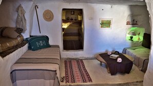 Ein gemütlicher, rustikaler Raum mit einem Bett, einem kleinen Tisch und einer Bank. Ein Durchgang zu einem anderen Raum, eine Wandregal mit Dekoration und ein gemustertes Teppichboden sind ebenfalls vorhanden.