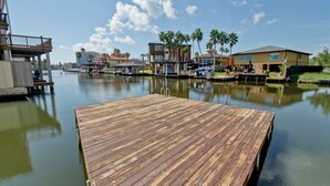 Un muelle de madera se extiende sobre aguas tranquilas, rodeado de edificios residenciales con balcones a la vista y palmeras.