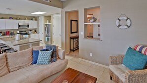 Una sala de estar con un sofá color beige, una mesa de centro de madera y una silla de mimbre con cojines de colores. Se ve una cocina con armarios blancos y un microondas al fondo.
