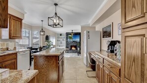 A spacious kitchen with wooden cabinets, a granite countertop, and a central island. There is a stone fireplace in the background, and a ceiling fan is visible.
