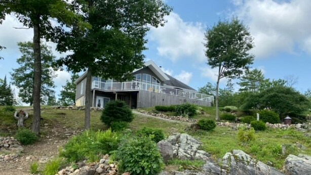 A modern house with a large deck, surrounded by a landscaped garden with rocks and trees. - Water and Beach Front Property 4 season on Ottawa River with Private Boat Launch (Deep River)
