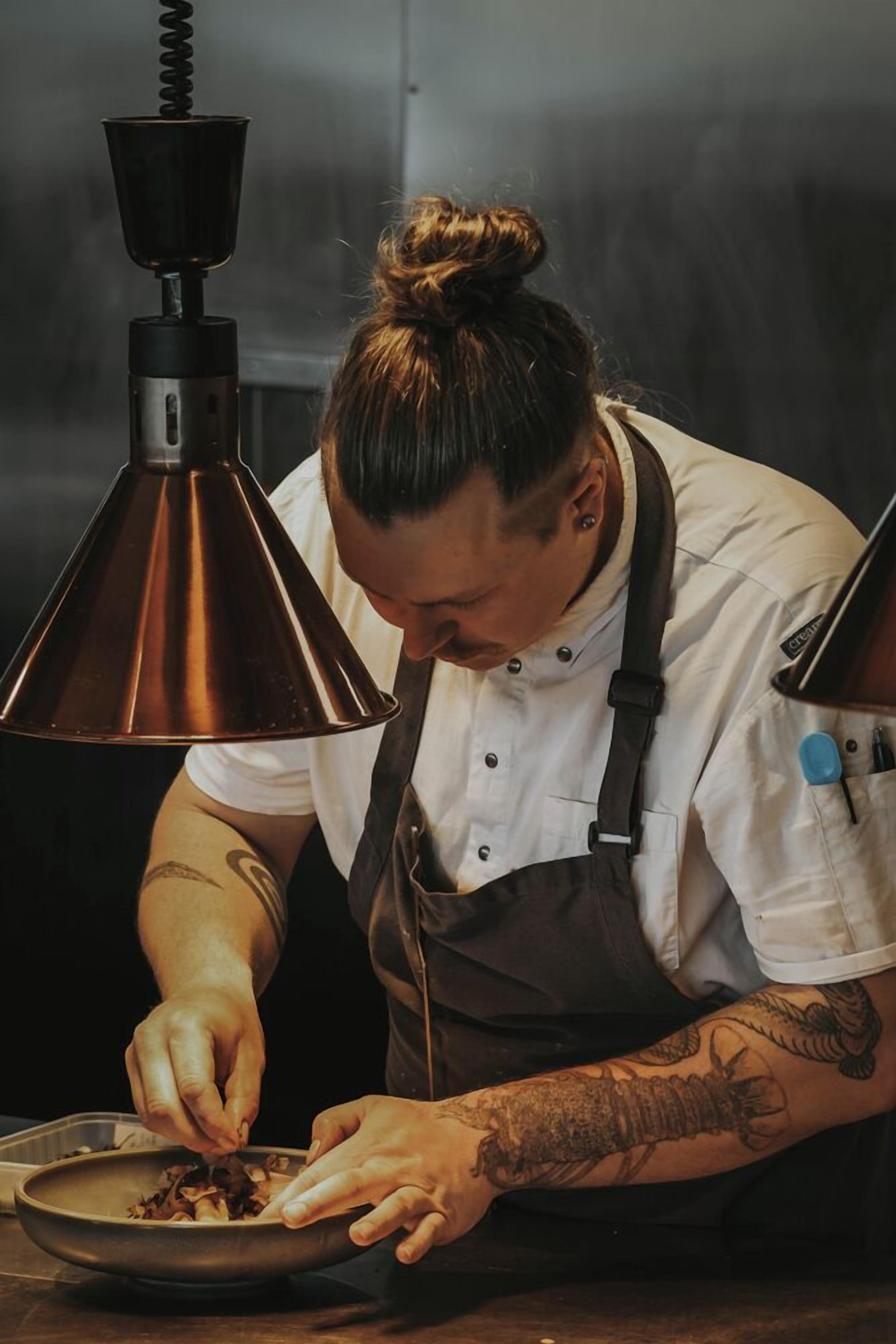 Un chef préparant des plats à la lumière d’une lampe de cuisine en cuivre, devant un évier en acier inoxydable.