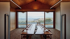 A conference room with a long table set for a meeting, large windows with a view of a lake and hills, and wooden ceiling beams.