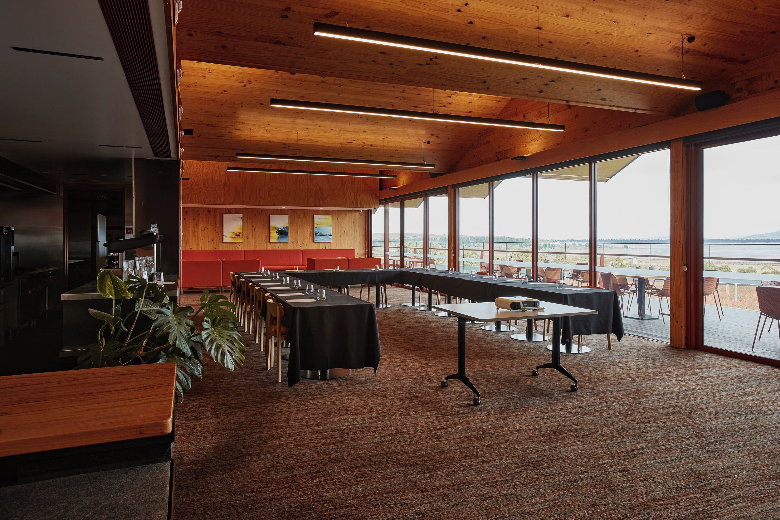 Une salle de conférence dotée de longues tables, d’un coin cuisine et de grandes fenêtres donnant sur l’extérieur.