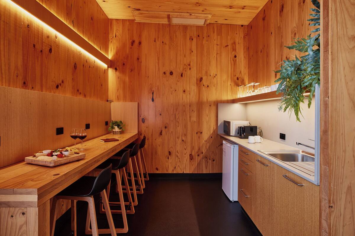 A compact kitchen with wooden walls, a long counter, bar stools, a sink, and a microwave.