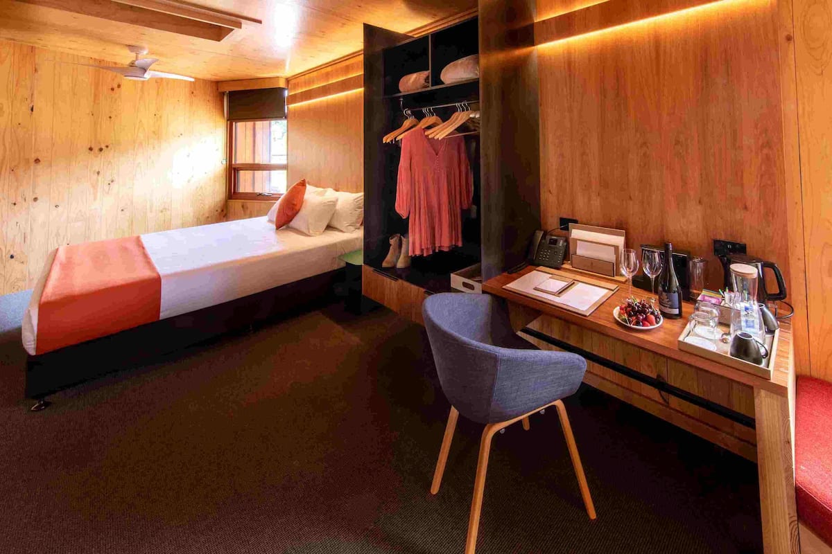 A wooden room with a bed, a desk, and a chair. There is a window with a view of greenery outside.