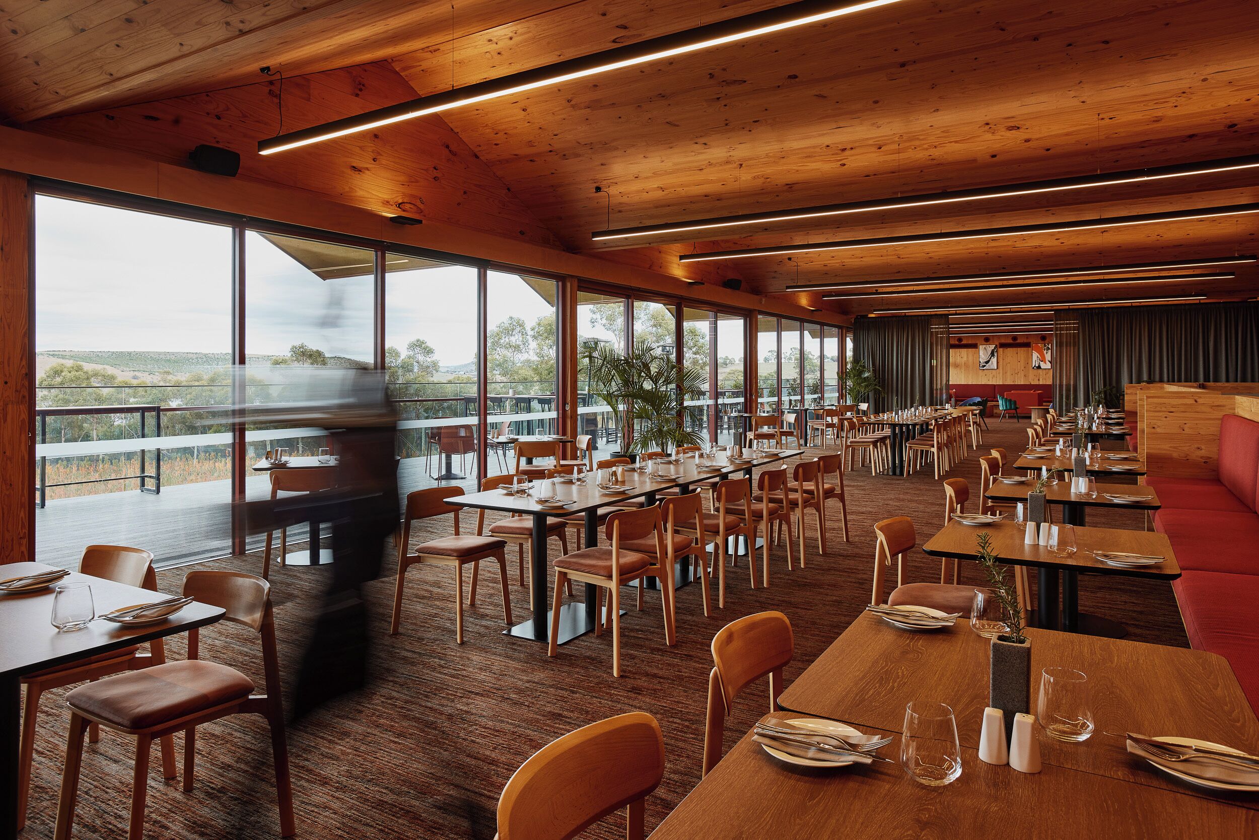 Un restaurante espacioso con mesas y sillas largas, ventanas amplias que ofrecen vistas al exterior y un techo de madera.