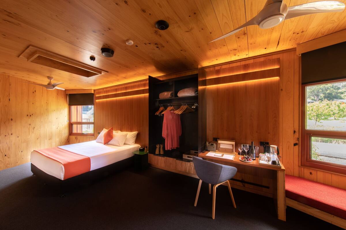 A wooden room with a bed, a desk, a chair, a wardrobe, and a window with a view.