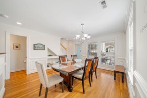 A dining area with a wooden table and chairs, a chandelier, and a view through the window.