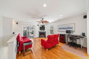 A spacious living room with a fireplace, a red sofa, a red armchair, a desk with a computer, and a ceiling fan.