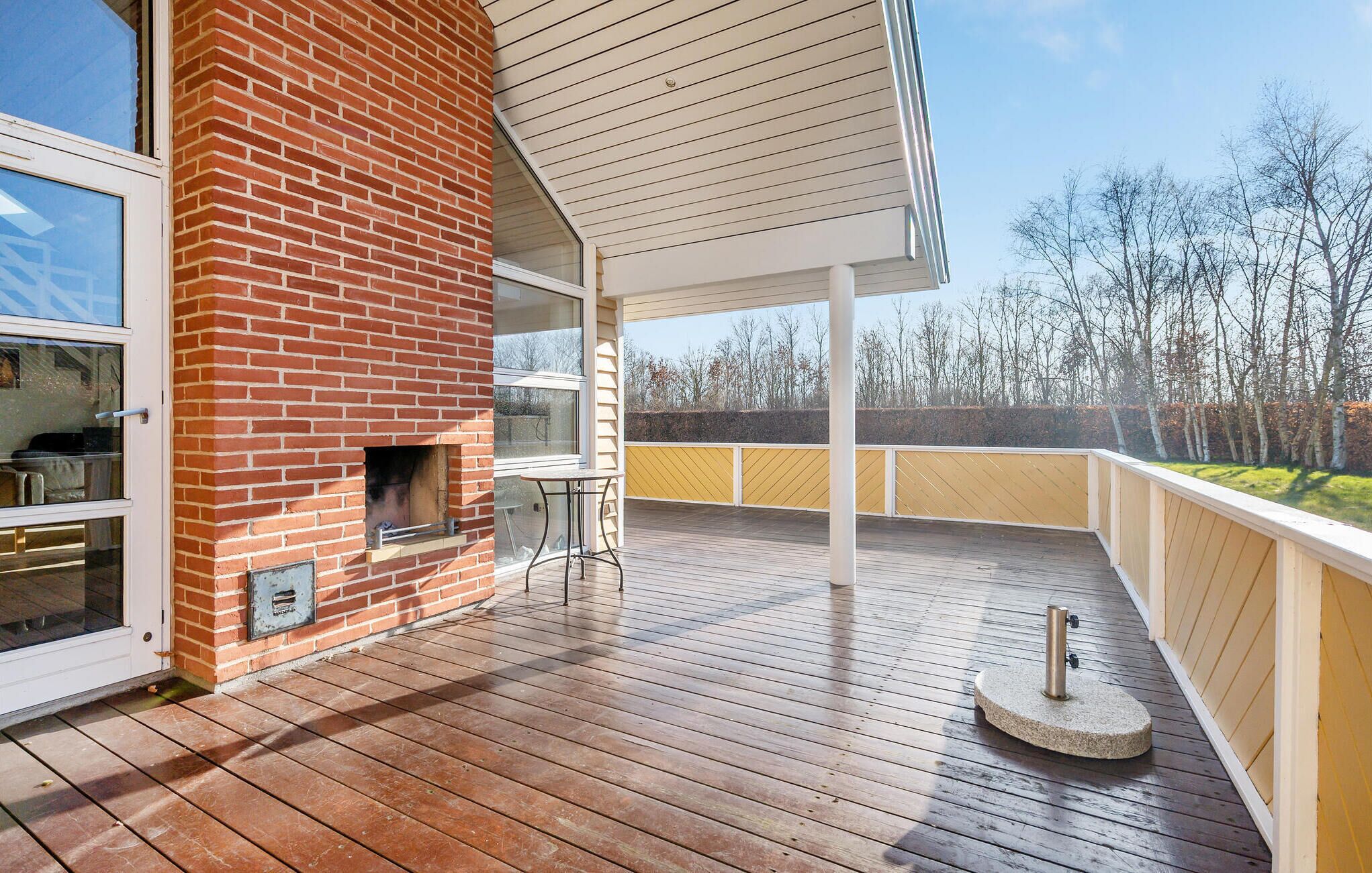 A wooden deck with a brick fireplace, outdoor seating, and a view of trees.