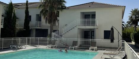 A swimming pool area with a tiled deck, lounge chairs, and a building with a balcony.